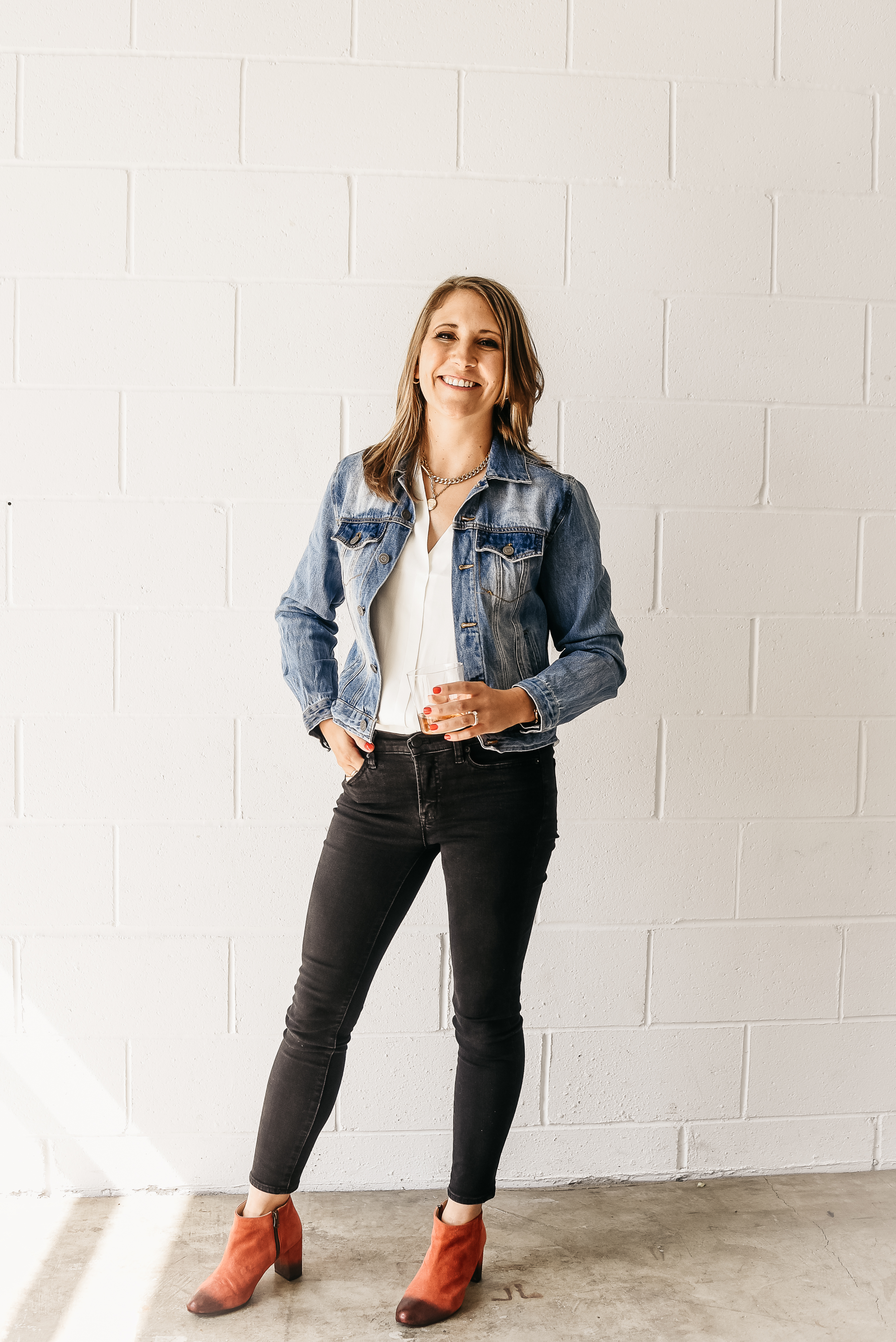 Photo of brand messaging strategist Annie Obergefell standing against a white brick wall holding a glass of bourbon and smiling