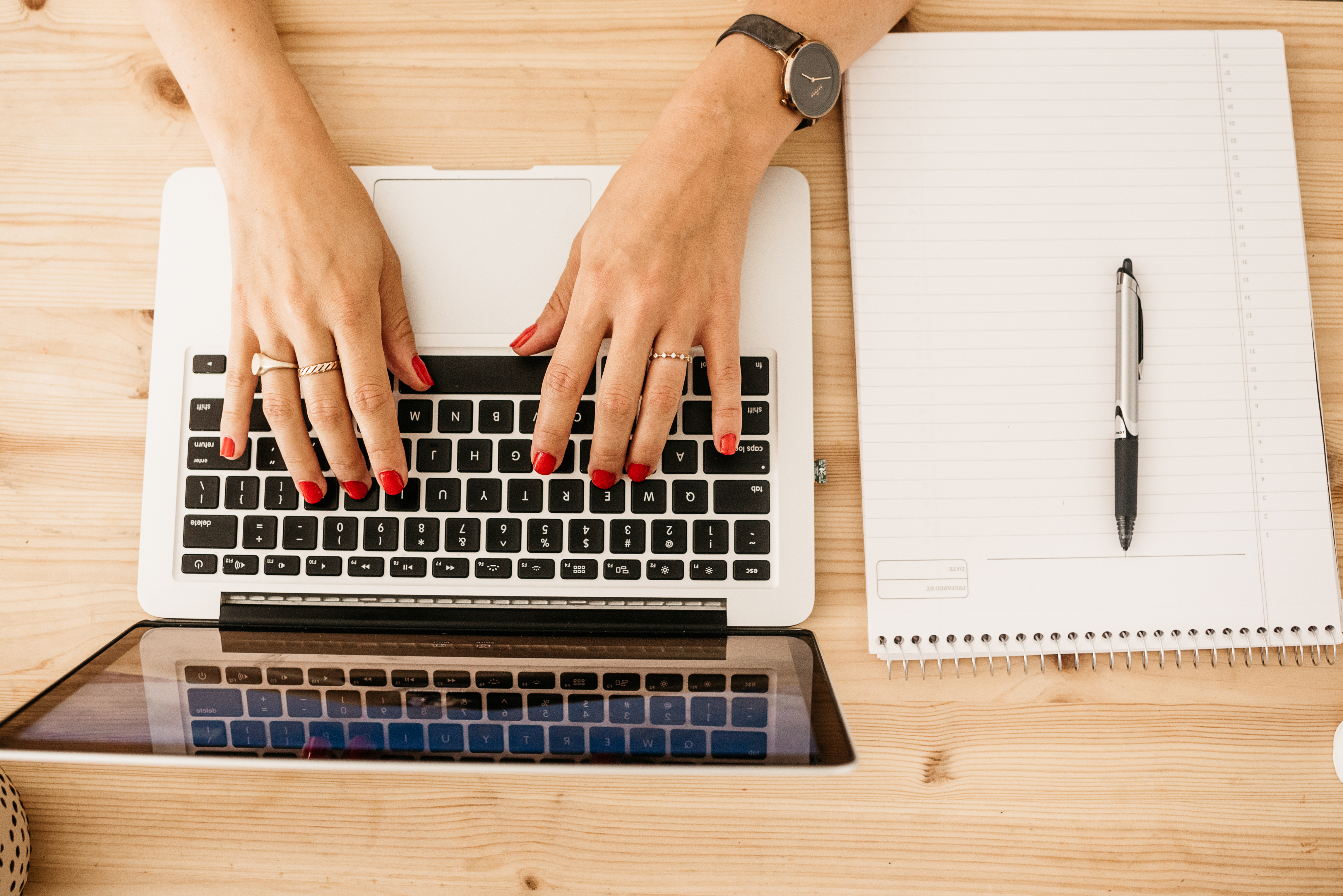 Image of an aerial view of a white woman's hands typing on a Mac keyboard, wearing red nail polish and gold rings, with a pen and notepad to her right