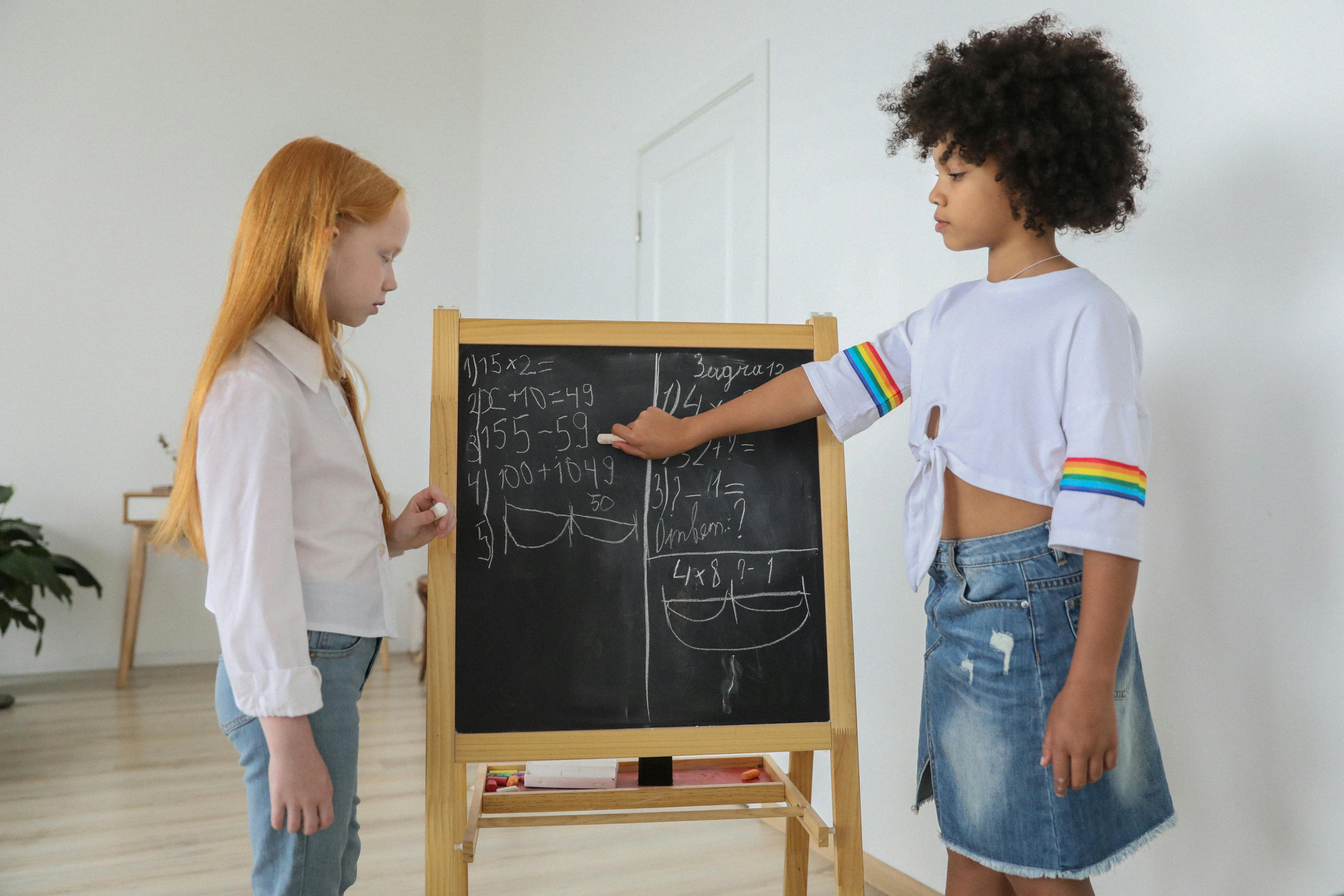 Image of a red headed girl and African American girl pointing to a blackground and solving a problem